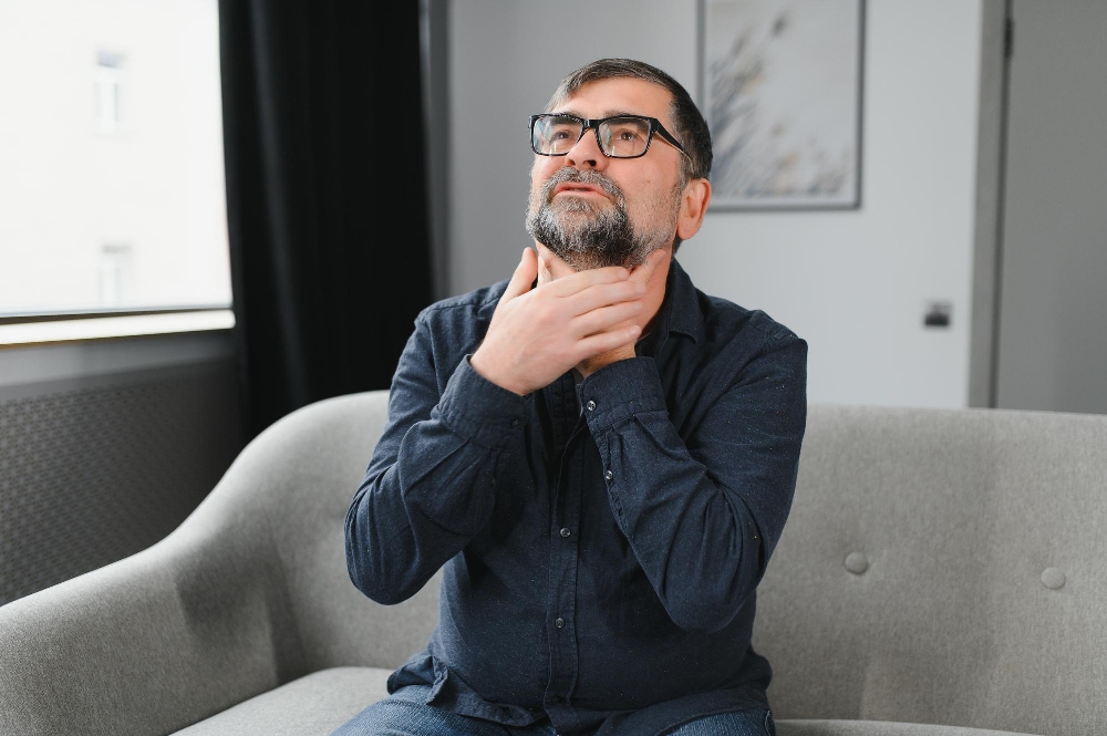 A man touching his throat while seated on a gray couch showing stroke dysphagia